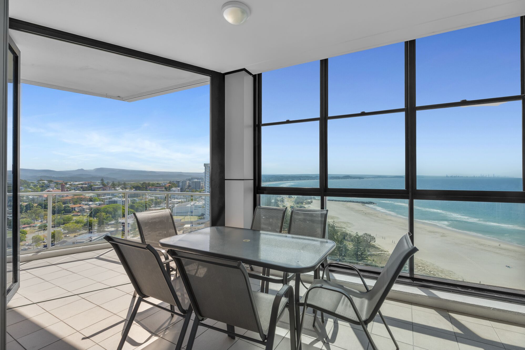 Un balcón con mesa y sillas de vidrio, con vistas a la playa y al paisaje urbano.
