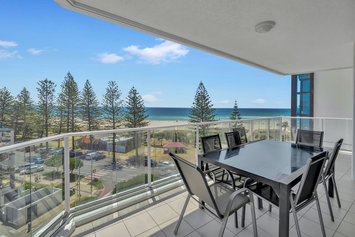 A balcony with a glass railing overlooking a beach and ocean, a table with chairs, and a clear sky.