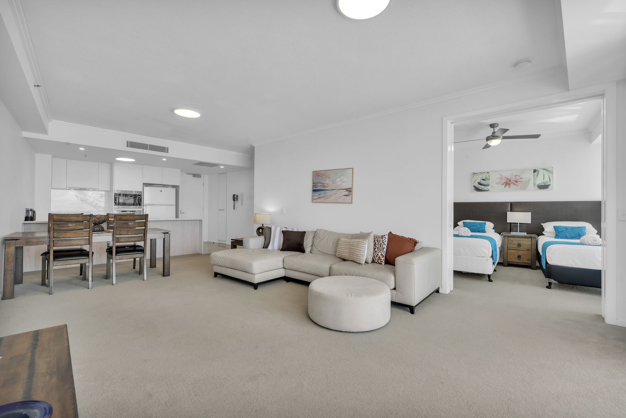 A modern living room with a sectional sofa, a round ottoman, a dining area with a wooden table and chairs, a kitchen with white cabinets, and a bedroom with two beds and a ceiling fan.