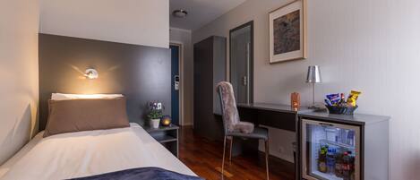 A modern hotel room with a bed, a desk, a chair, a mini-fridge, and a mirror.