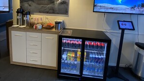 Un espace d’accueil d’hôtel comprenant un réfrigérateur rempli de boissons, une machine à café et un grand tableau représentant un coucher de soleil sur des montagnes.