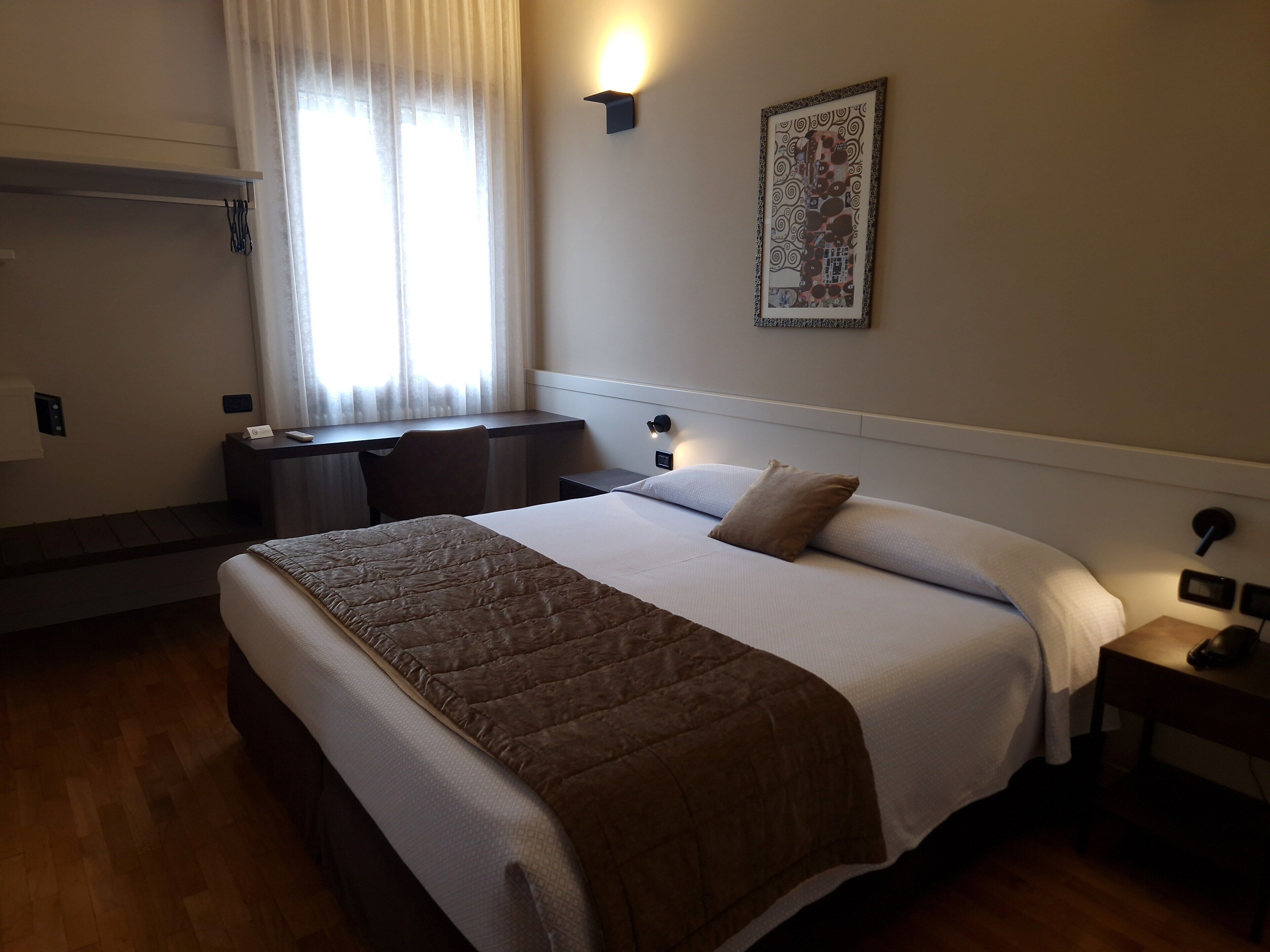 A hotel room with a bed, a desk, a chair, a nightstand, and a wall-mounted light.