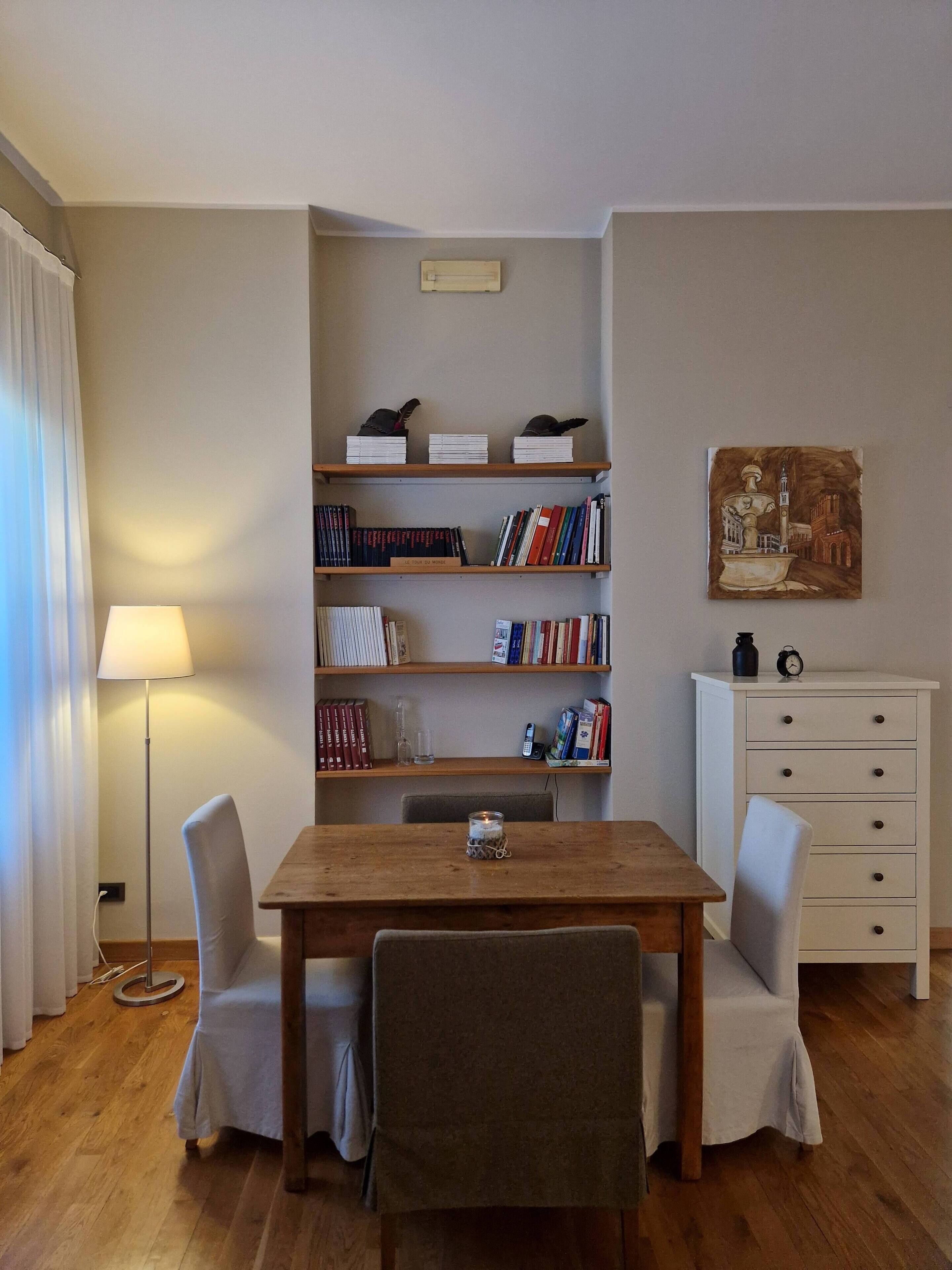 Una habitación acogedora con una mesa de madera, dos sillas, una estantería, un armario blanco y una lámpara.