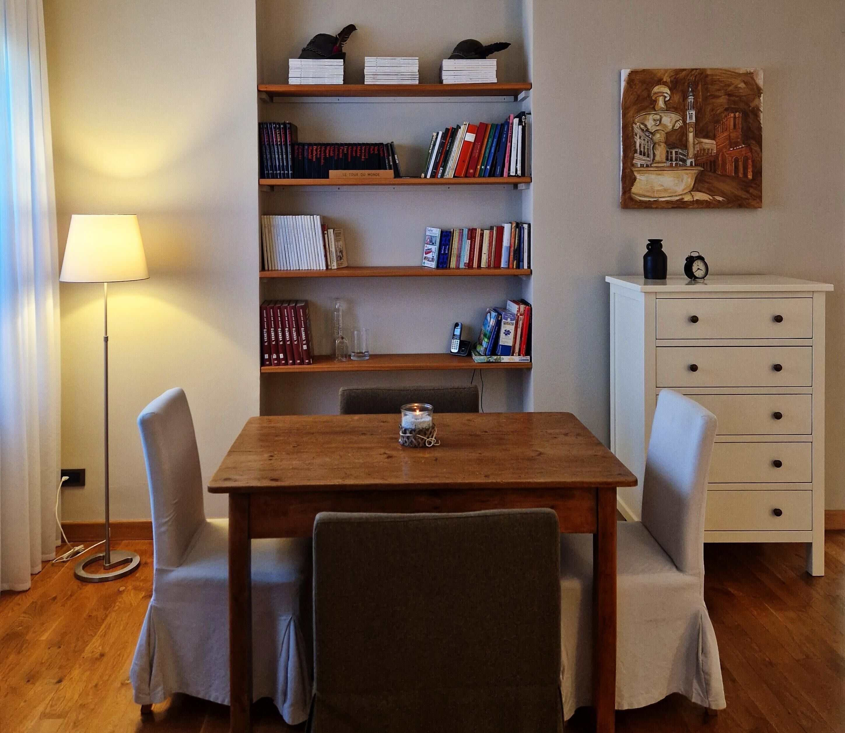 A room with a wooden table, two chairs, a bookshelf, a white dresser, and a painting on the wall.