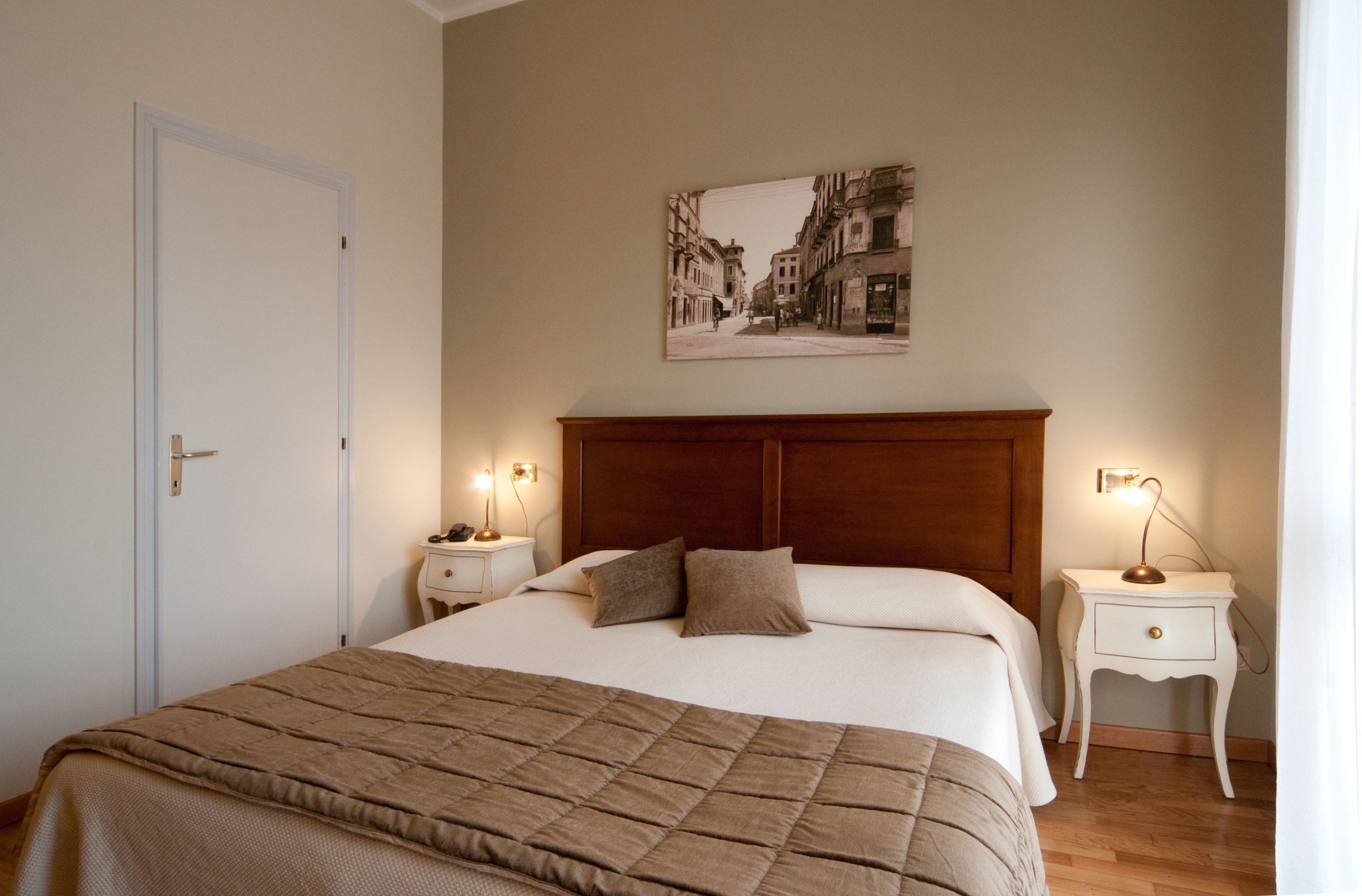 Un letto rifatto con un copriletto marrone, due comodini con lampade e un quadro appeso al muro raffigurante una scena di strada.