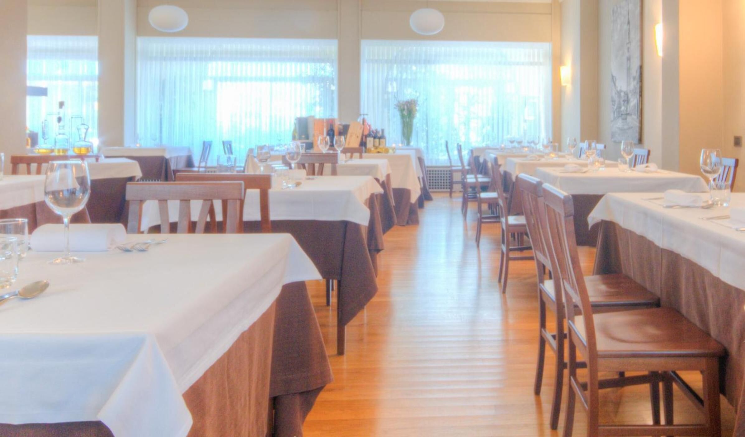 Een restaurant met meerdere tafels, elk gedekt met witte tafellnen en bestemd voor servies en wijnglazen.