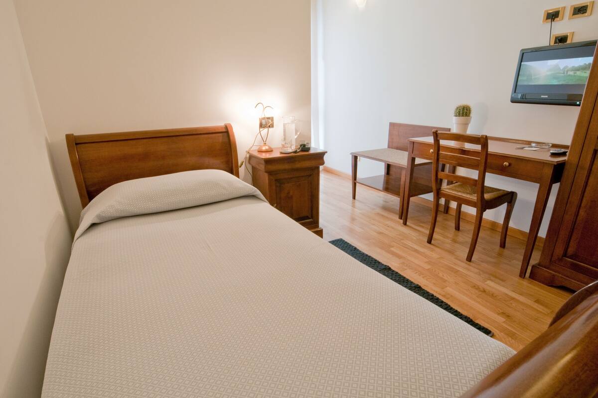 A bedroom with a bed, a desk, a chair, a nightstand, and a television.