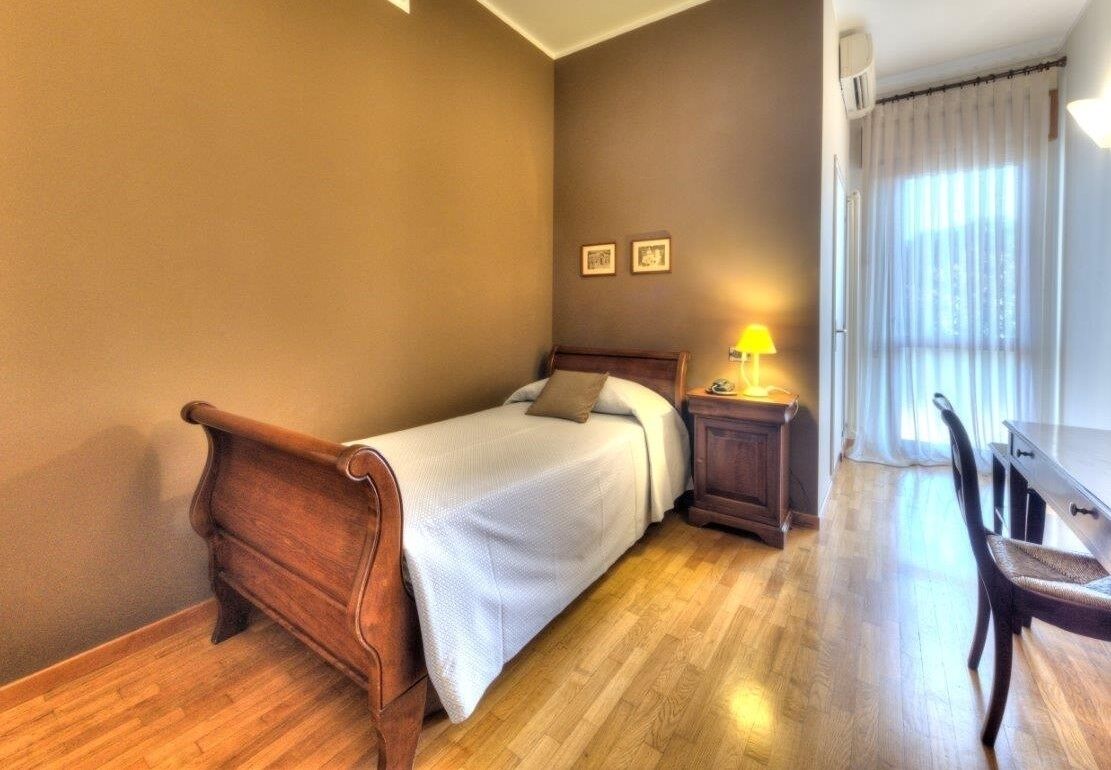A bedroom with a wooden bed, a nightstand with a lamp, a chair, and a window with curtains.