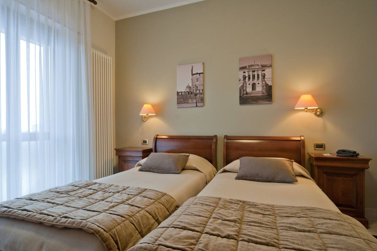 A hotel room with two beds, a window with sheer curtains, wall-mounted lamps, and two framed pictures.