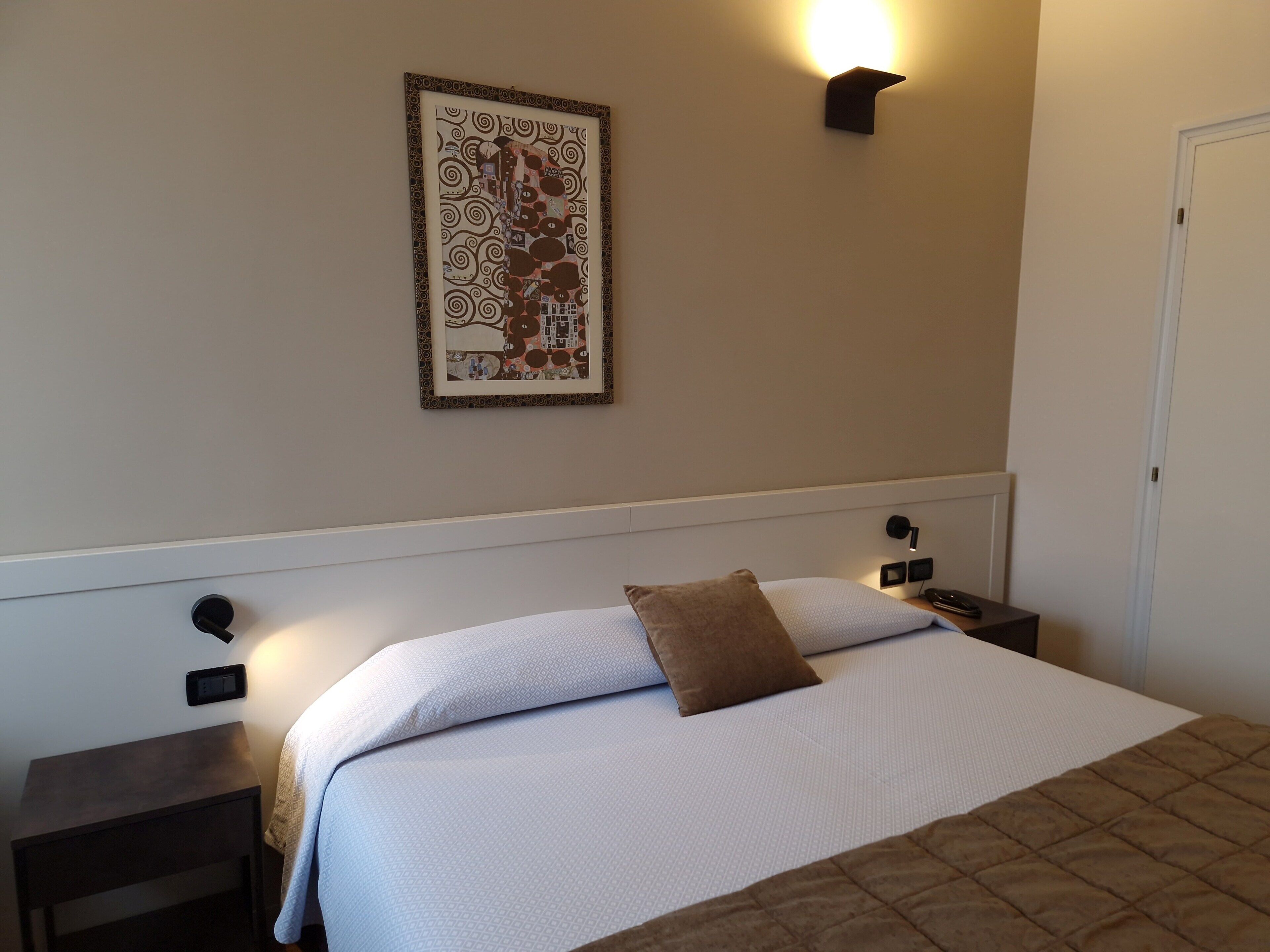 A neatly made bed with a patterned pillow, a framed artwork on the wall, and a wall-mounted light fixture.