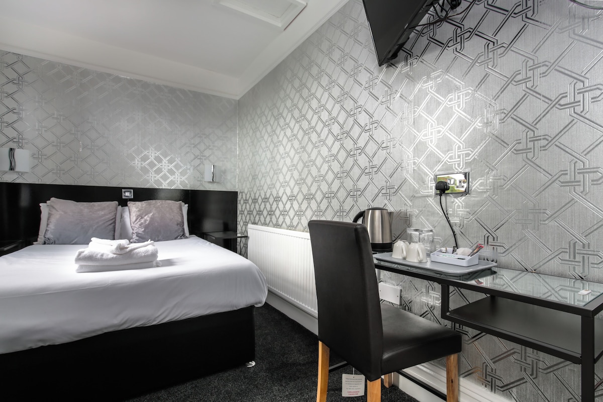 A hotel room with a bed, a desk, and a chair. There is a radiator, a kettle, and a small table with cups and a phone.