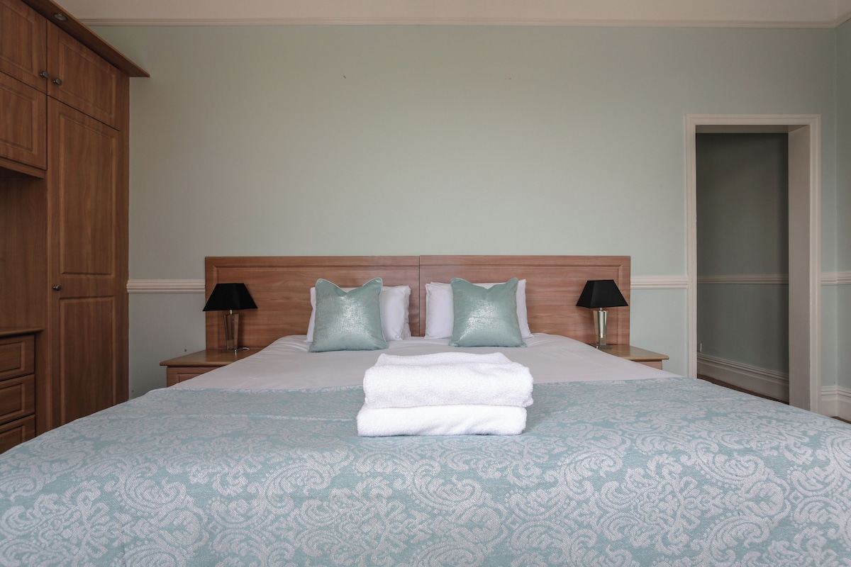 A neatly made bed with a patterned bedspread and pillows, a wooden headboard, two bedside lamps, and a wooden wardrobe.