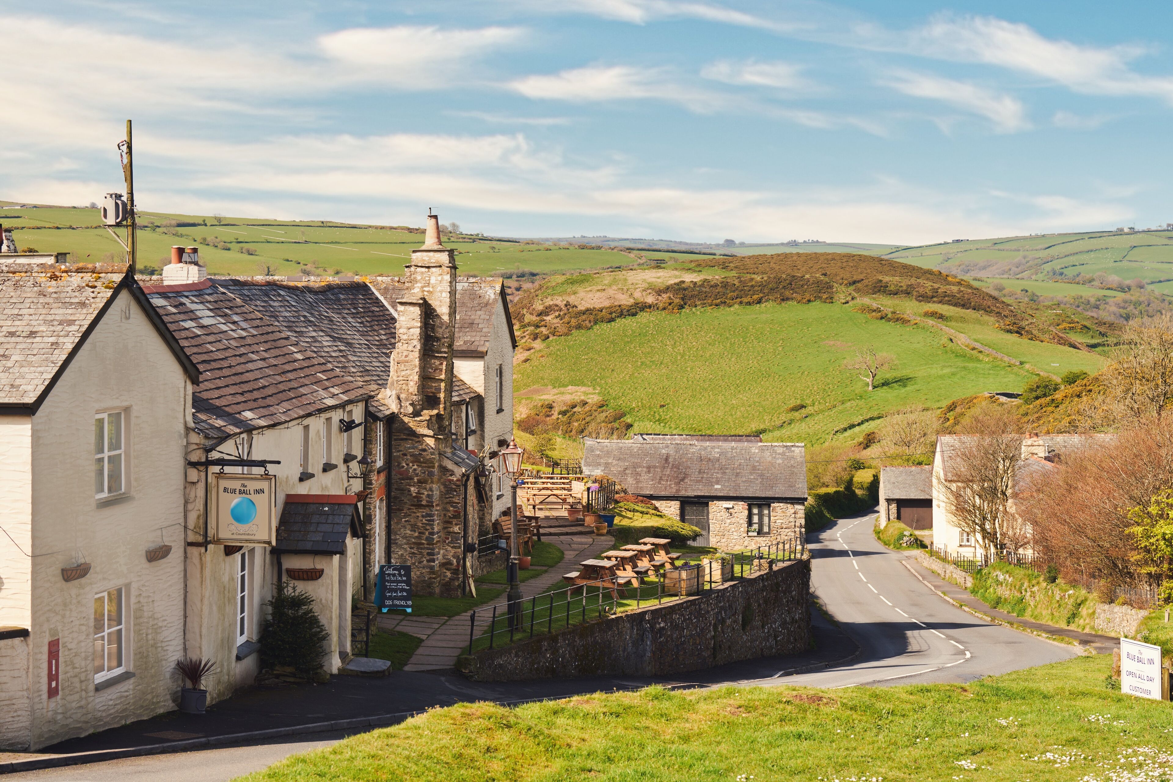 A traditional stone building with a sign for the Blue Ball Inn, a paved road, and rolling green hills in the background.