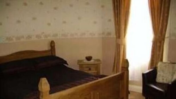 A wooden bed with a headboard, a bedside table, and a chair in a room with patterned wallpaper. - Boars Head Hotel (Middlewich)