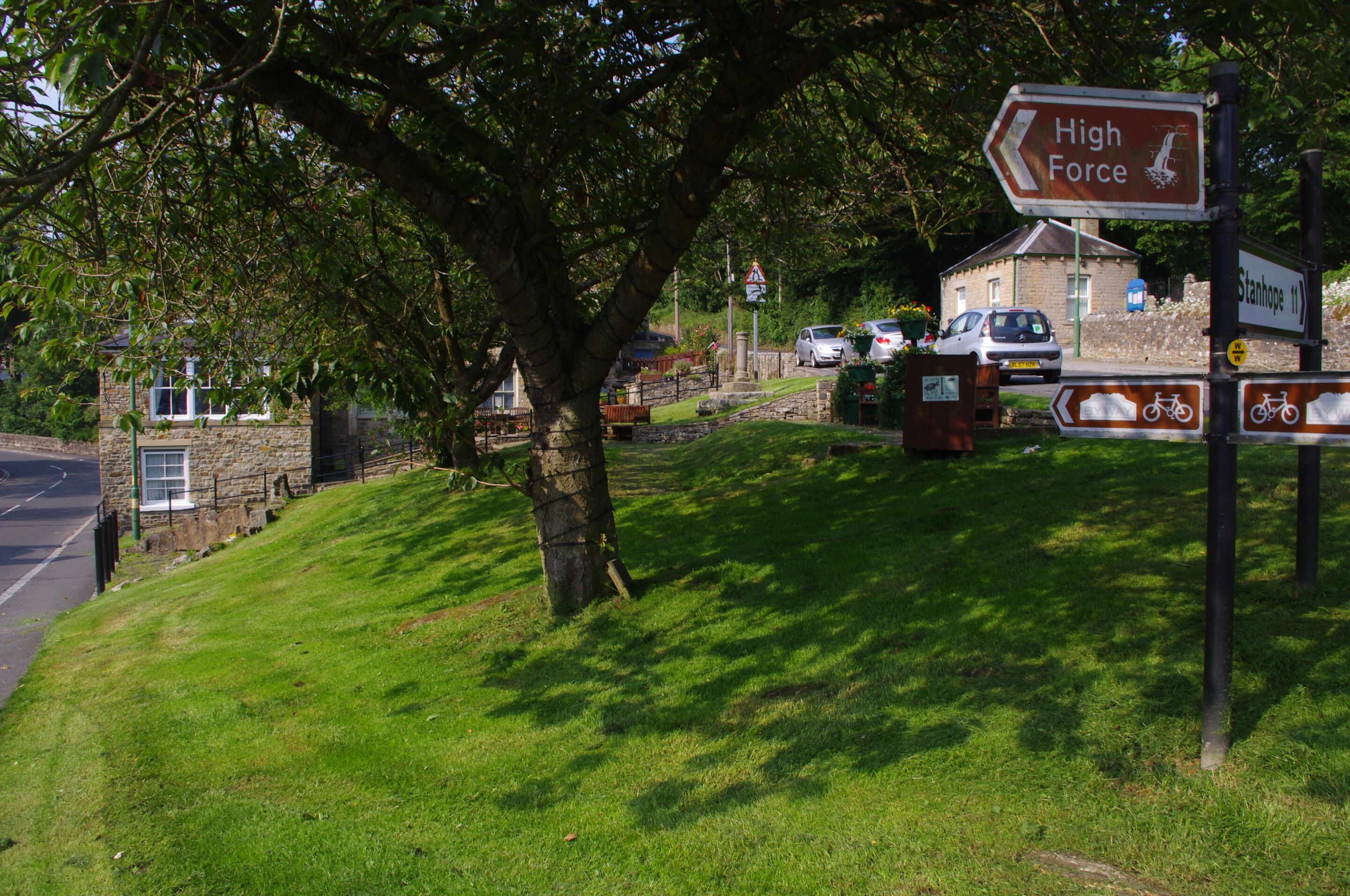 A signpost with directions to High Force and Stanhope, a stone building, and a grassy area with a tree.