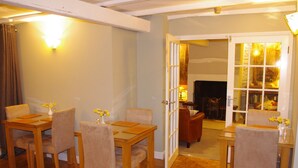 A dining area with wooden tables and chairs, a vase with flowers, and a doorway leading to another room with a fireplace. - Brunswick House (Barnard Castle)