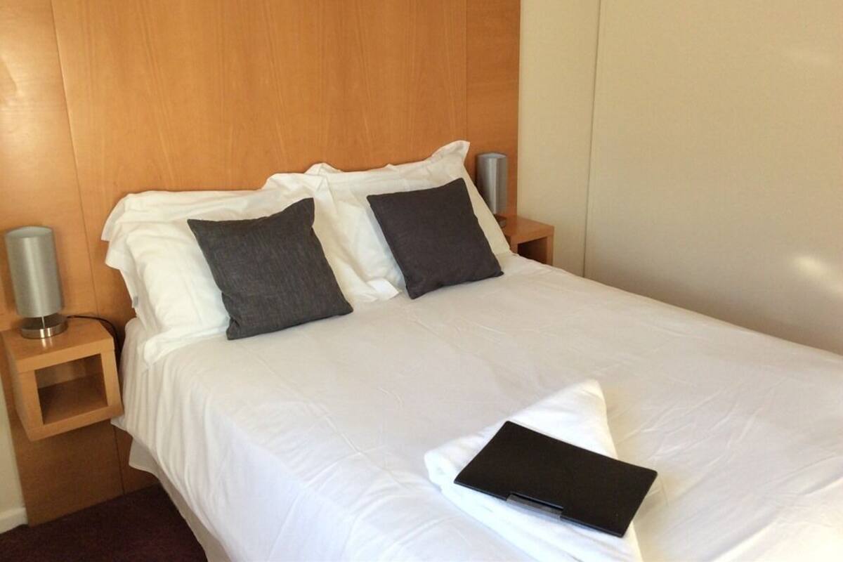 A neatly made bed with white linens and two dark pillows, a bedside table with a lamp, and a black object on the bed.