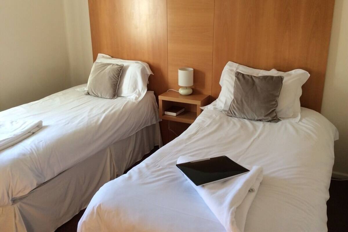 A hotel room with two single beds, each with white linens and pillows. There is a bedside table with a lamp and a book.