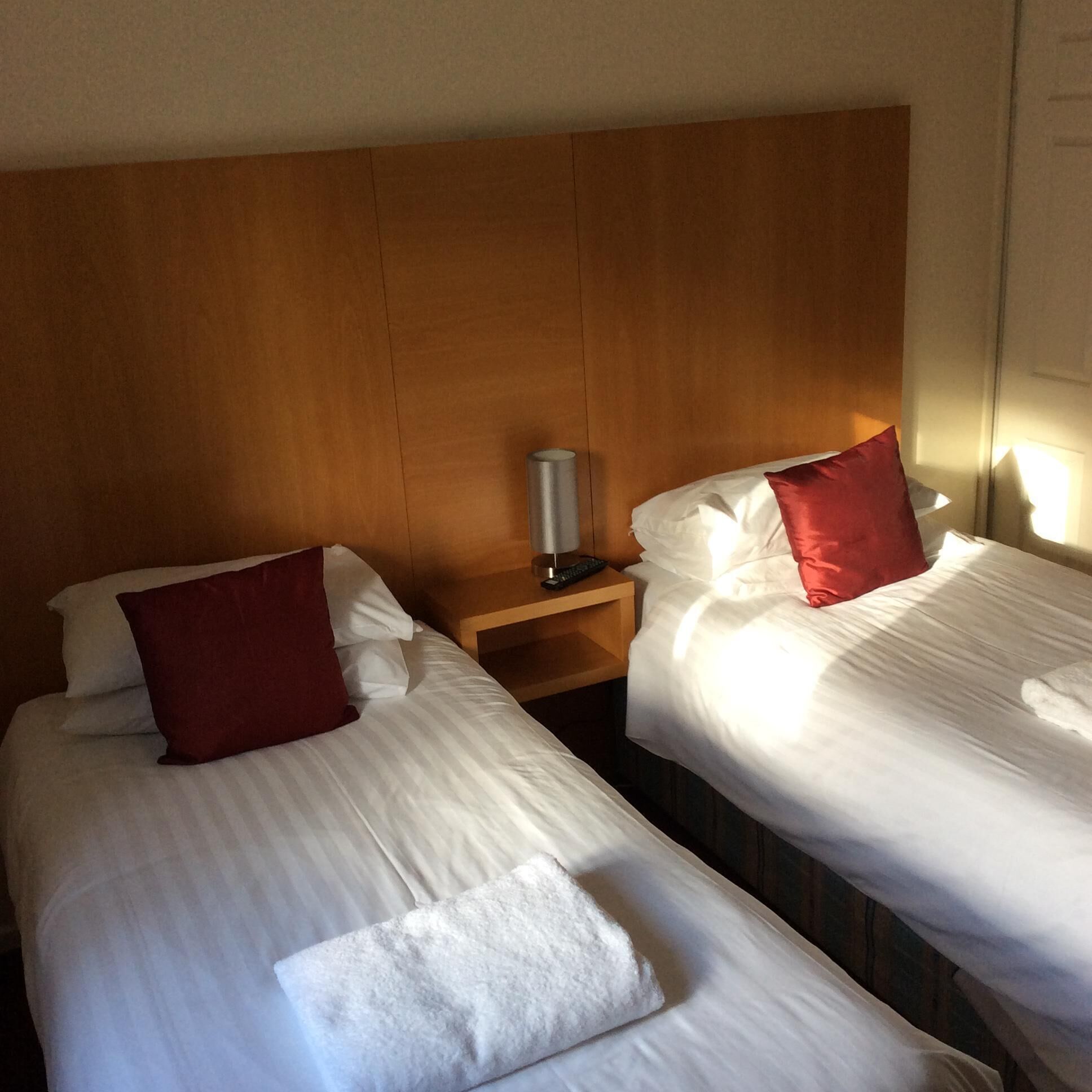 Two single beds with white linens and red pillows, a wooden headboard, a bedside table with a lamp, and a nightstand with a remote control.