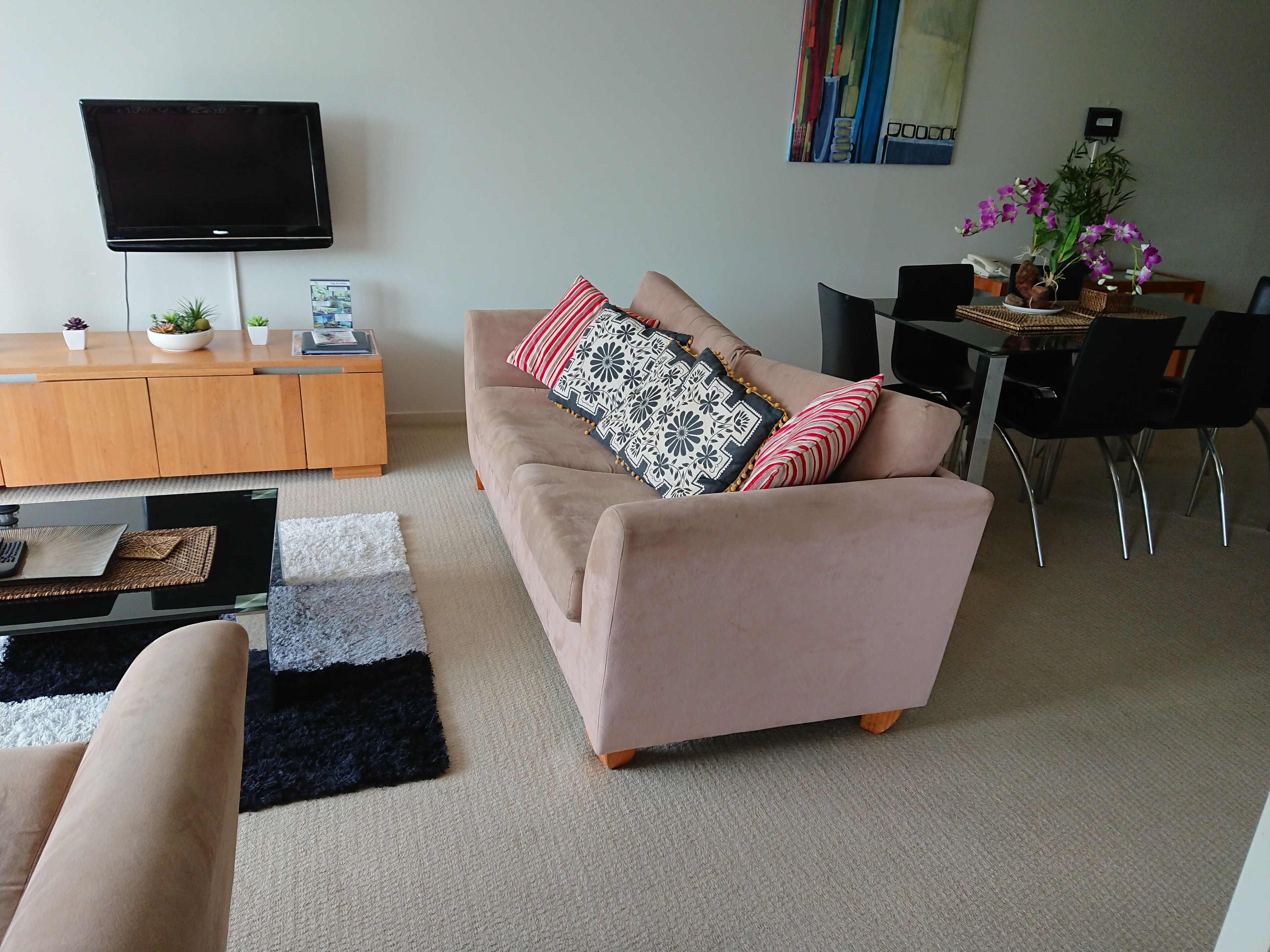 Uma sala de estar com sofá bege, TV de tela plana na parede, uma mesa pequena com planta em vaso e um espaço de jantar com uma cadeira.