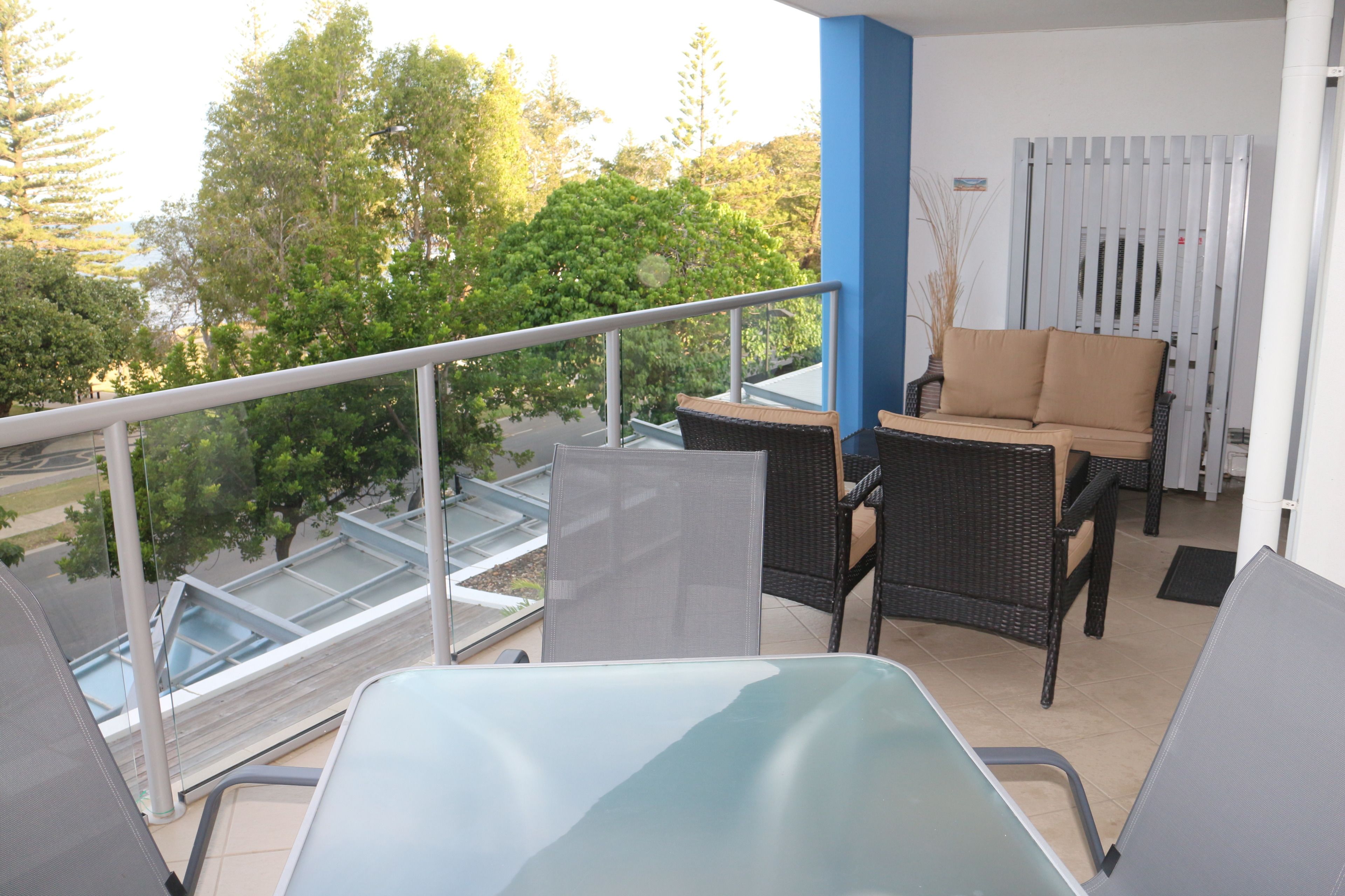A balcony with wicker furniture and a glass table, overlooking a green area with trees.