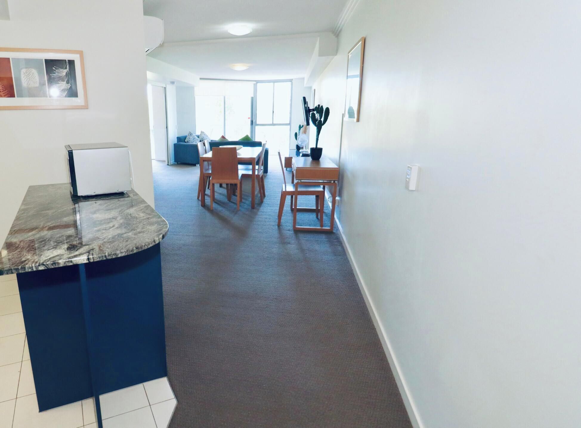 A modern kitchen with a marble countertop, a dining area with wooden chairs and a table, and a hallway leading to a living room.