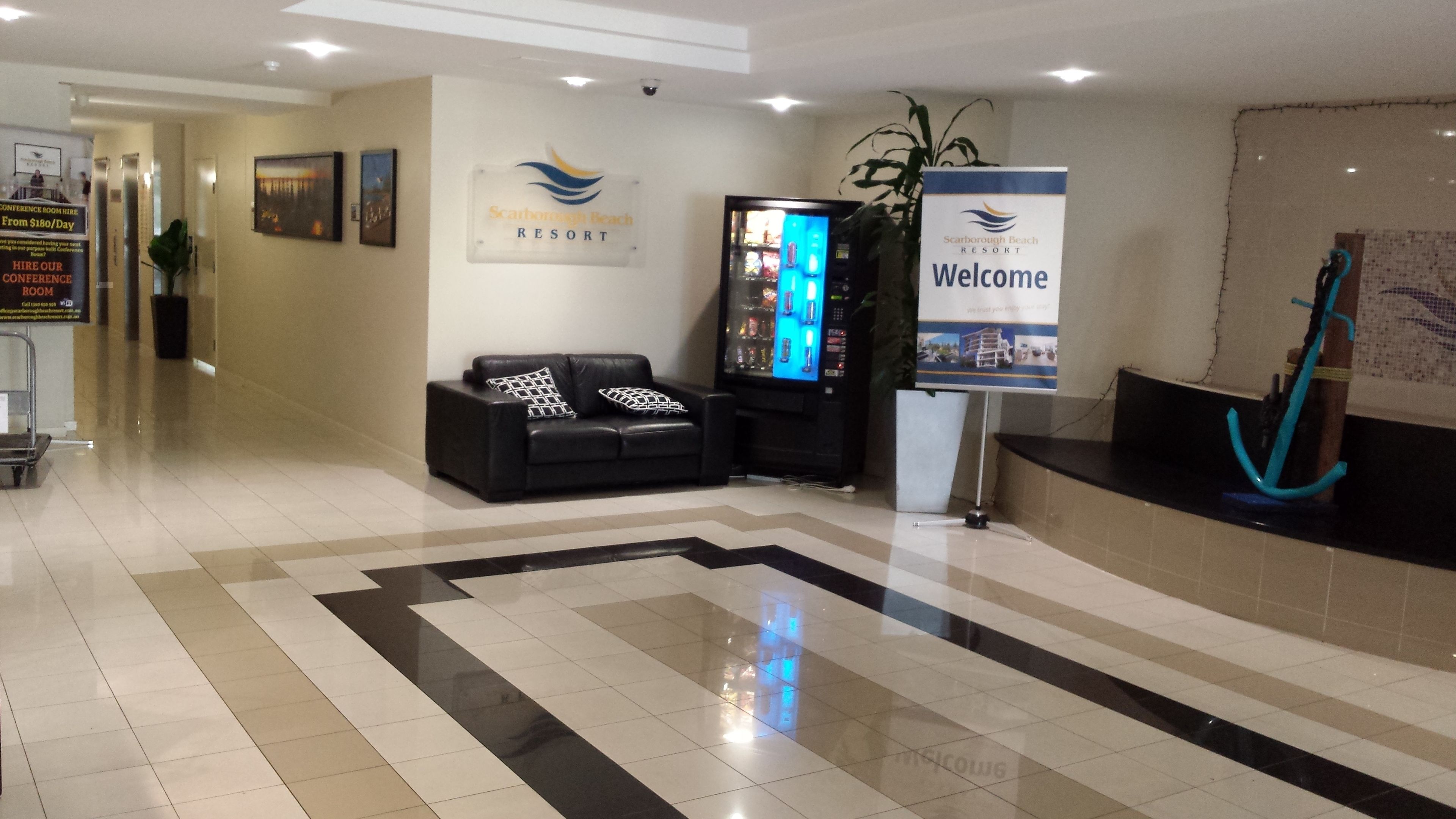 A reception area with a black leather sofa, a welcome sign, a vending machine, and a plant.