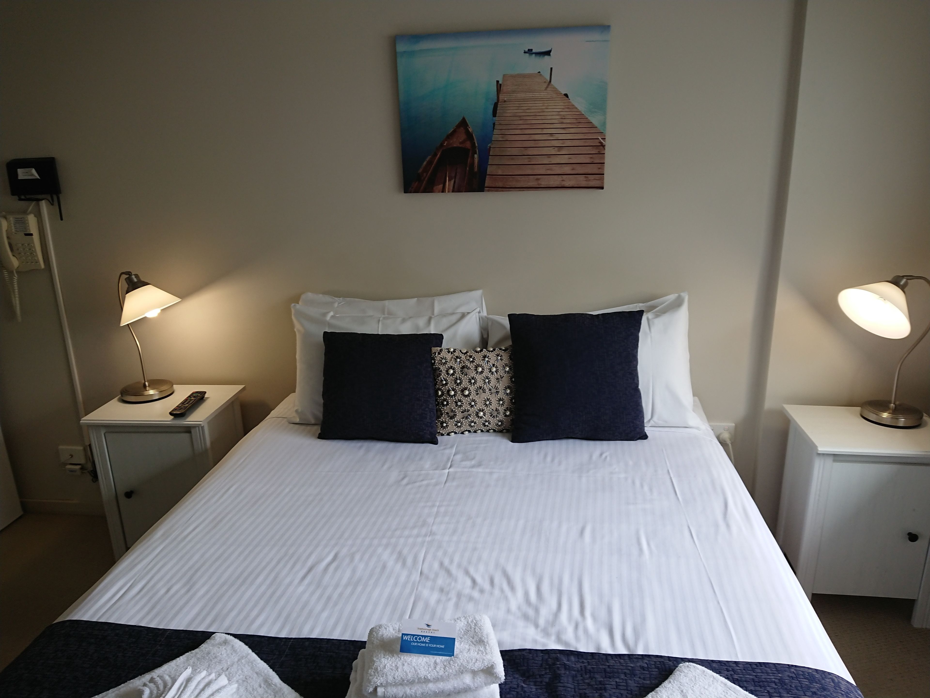 A neatly made bed with white linens, two bedside lamps, a painting of a pier, and a welcome towel on the bed.