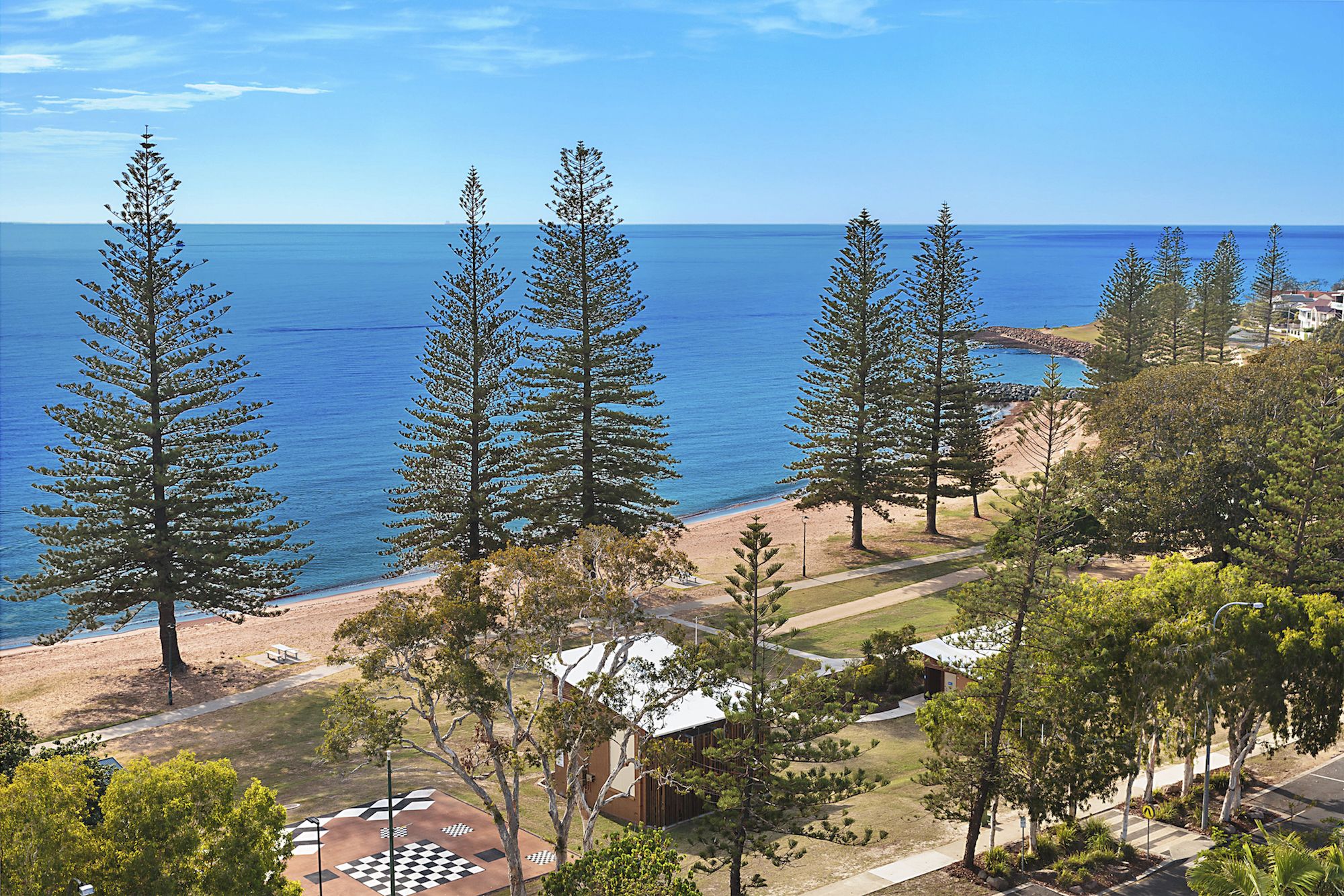 Una vista costiera con alti pini, una spiaggia e un edificio con un tetto dalla forma geometrica particolare.