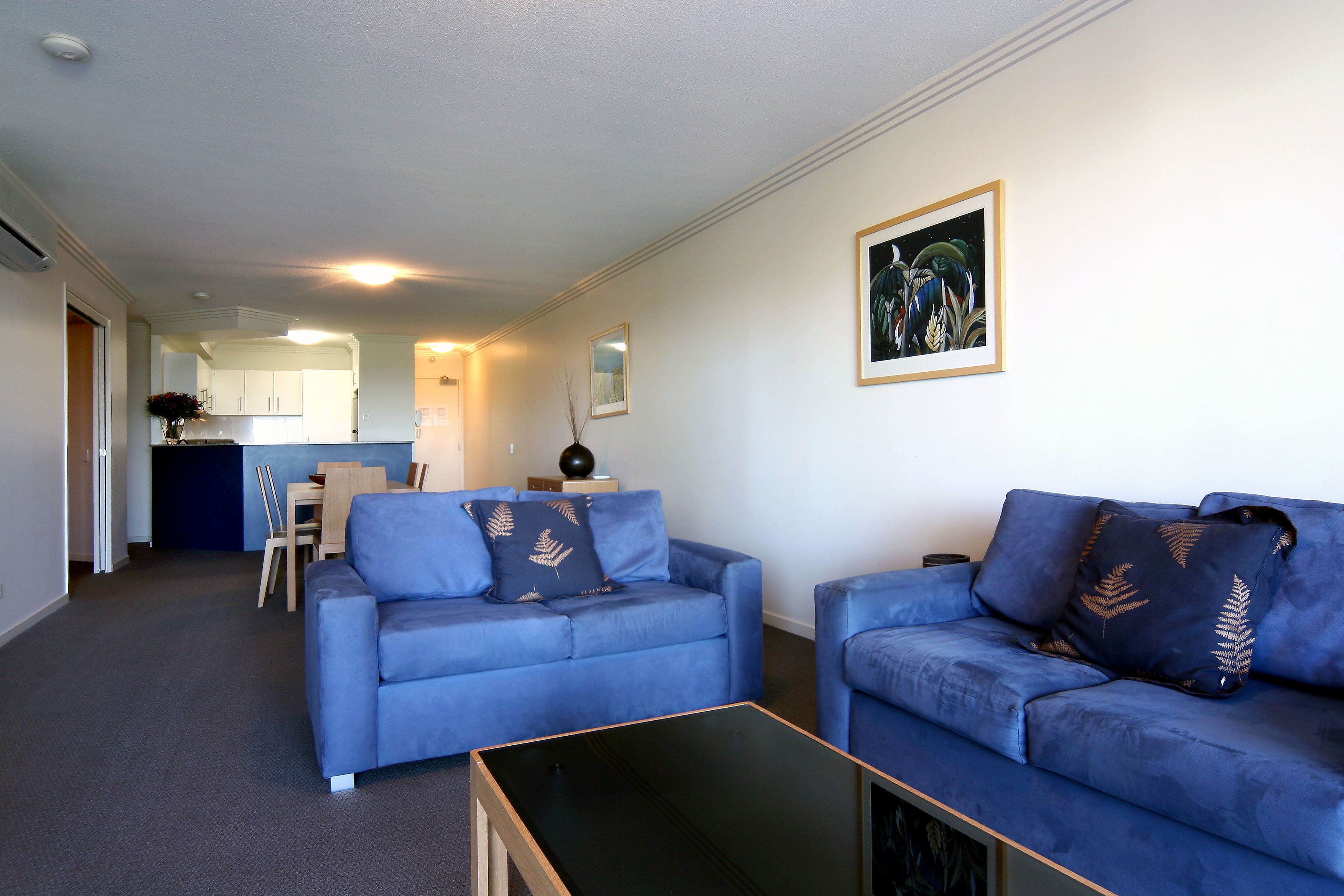 A modern living room with two blue sofas, a black coffee table, and a dining area in the background.