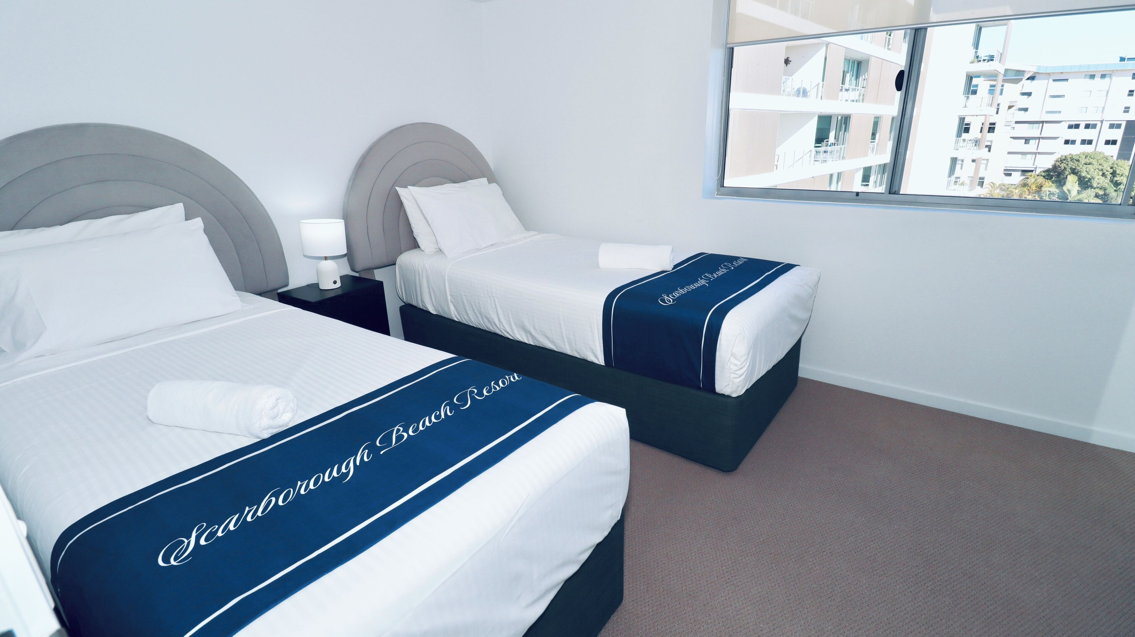 A hotel room with two beds, each with a headboard and a bedside table with a lamp. The room has a large window with a view of buildings.