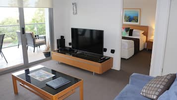 A modern living room with a flat-screen TV, a glass-top coffee table, a sofa, and a balcony with wicker chairs.