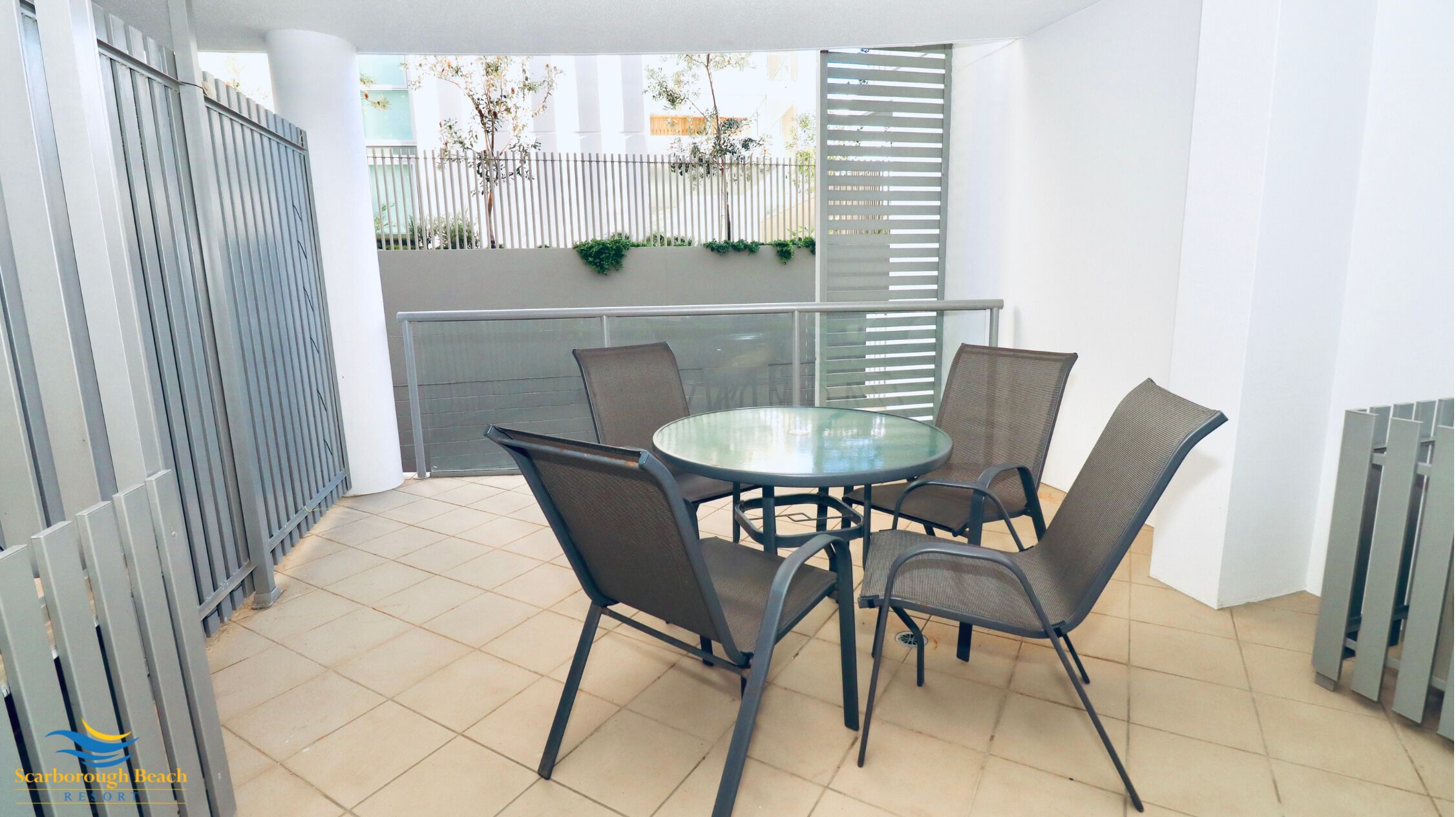 Een terras met een ronde glazen tafel en vier stoelen, een metalen hek en uitzicht op een gebouw met balkon.