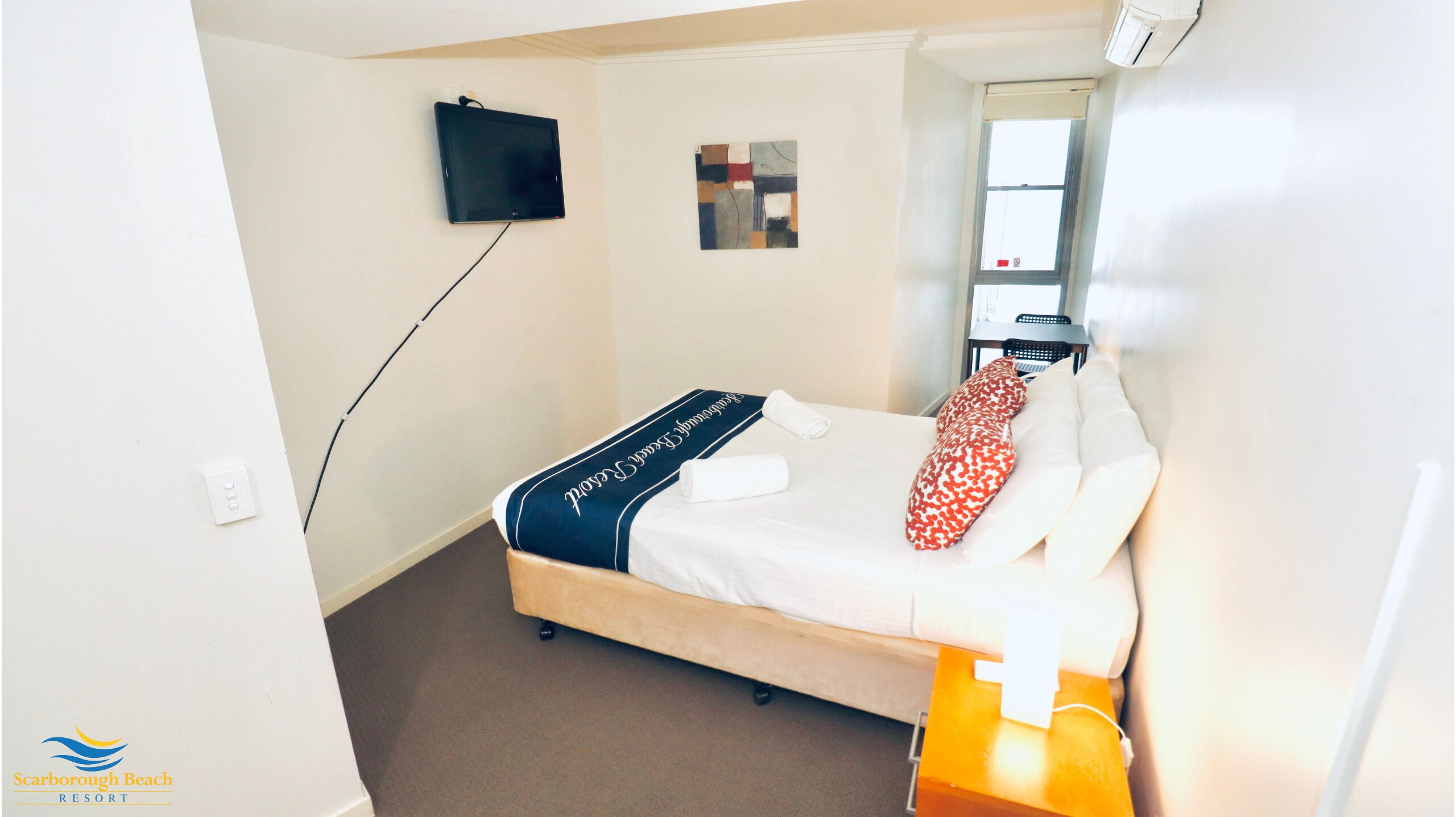 Hotellihuone, jossa on sänky, seinälle asennettu taulutelevisio, pieni taideteos, ikkuna ja yöpöytä, jossa on lamppu.