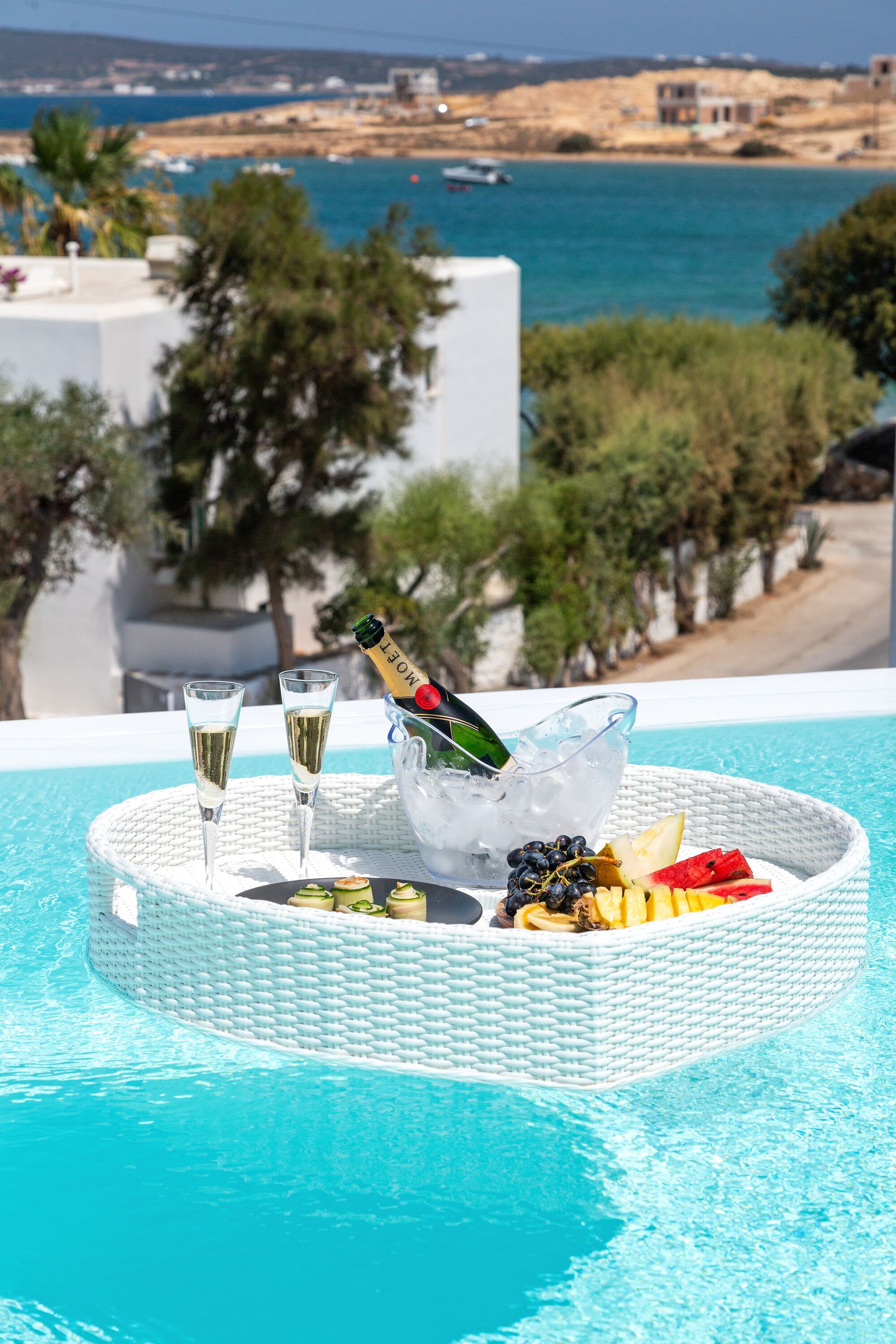 A poolside setting with a tray containing two glasses of champagne, a bottle of champagne, and a bowl of fruit.