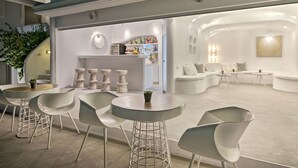 Zona de bar moderna con interior blanco, mesas redondas y taburetes. Hay un espacio de descanso con sofás y una barra repleta de botellas.