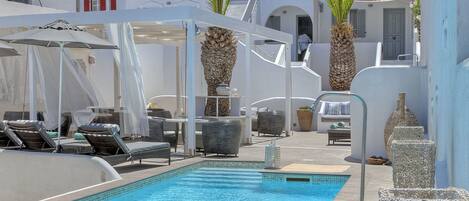 Zona de piscina con sillones de descanso, sombrillas y edificios blancos con balcones.