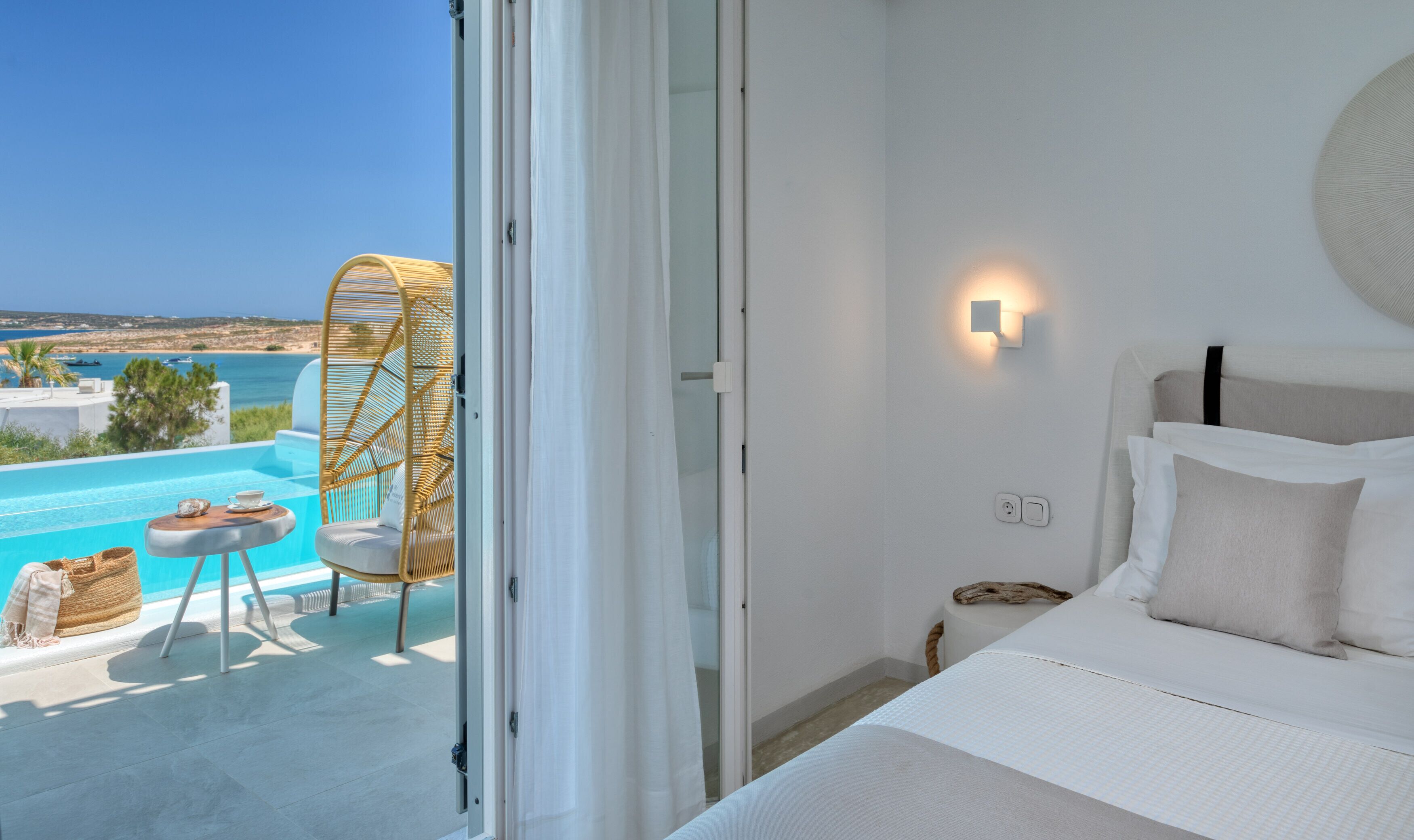 A bedroom with a view of a pool and the sea, a wicker chair, a bedside table with a lamp, and a bed with pillows.