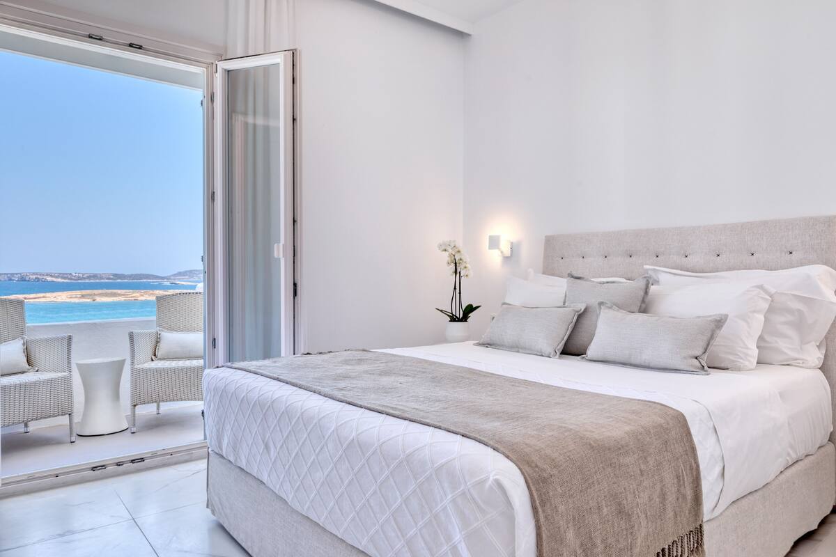 A bedroom with a large bed, a view of the beach, and a balcony with wicker furniture.