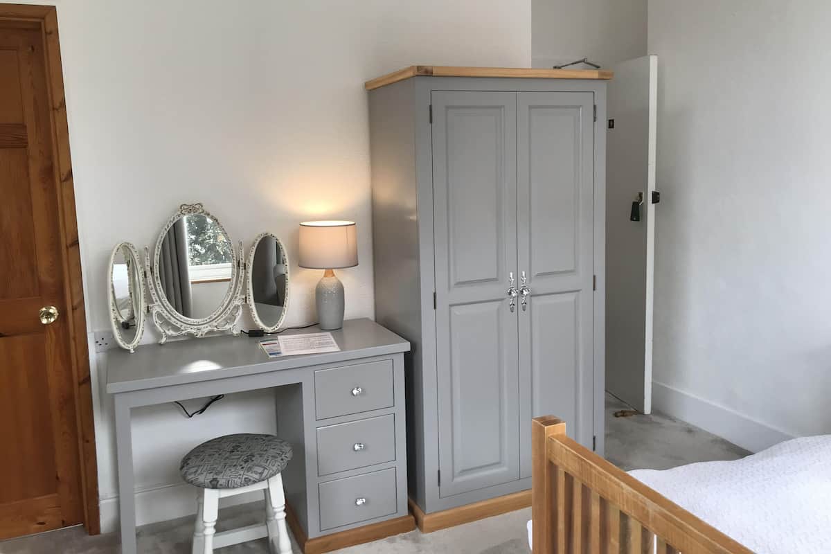 A bedroom with a wooden bed, a grey wardrobe, a vanity with a mirror and lamp, and a small stool.