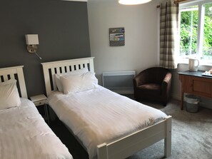 A bedroom with two single beds, a desk with a chair, a radiator, and a window with curtains. - Cornerways (Ventnor)