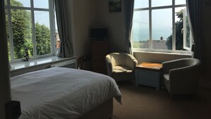 A hotel room with a bed, two armchairs, a small table, and large windows offering a view of the outdoors.