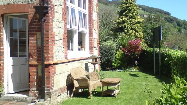 A brick house with a white door and windows, a small garden with a lawn and a chair, and a tall tree with red flowers.