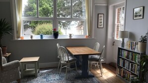 A cozy room with a wooden table and chairs, a bookshelf, a window with a view of greenery, and a patterned rug on the floor.