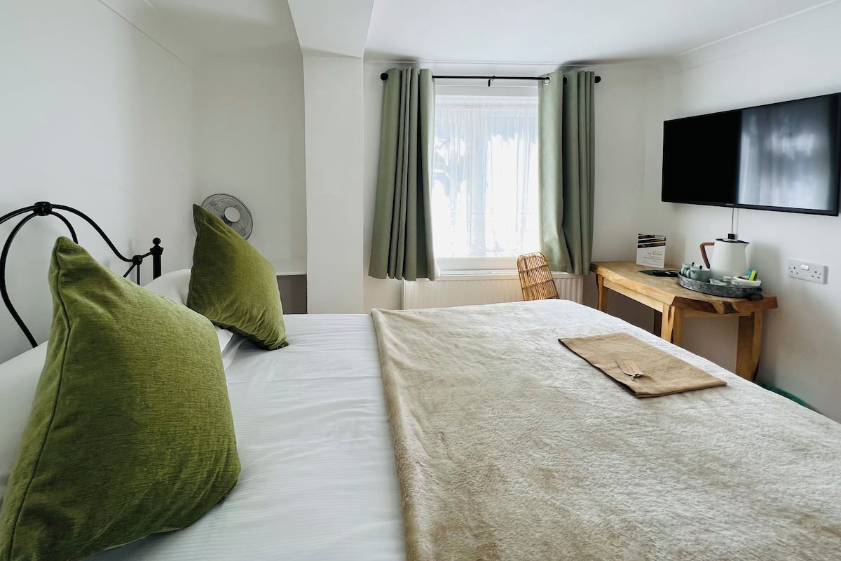A bedroom with a bed, two green pillows, a bedside table with a lamp, a chair, and a TV on the wall.
