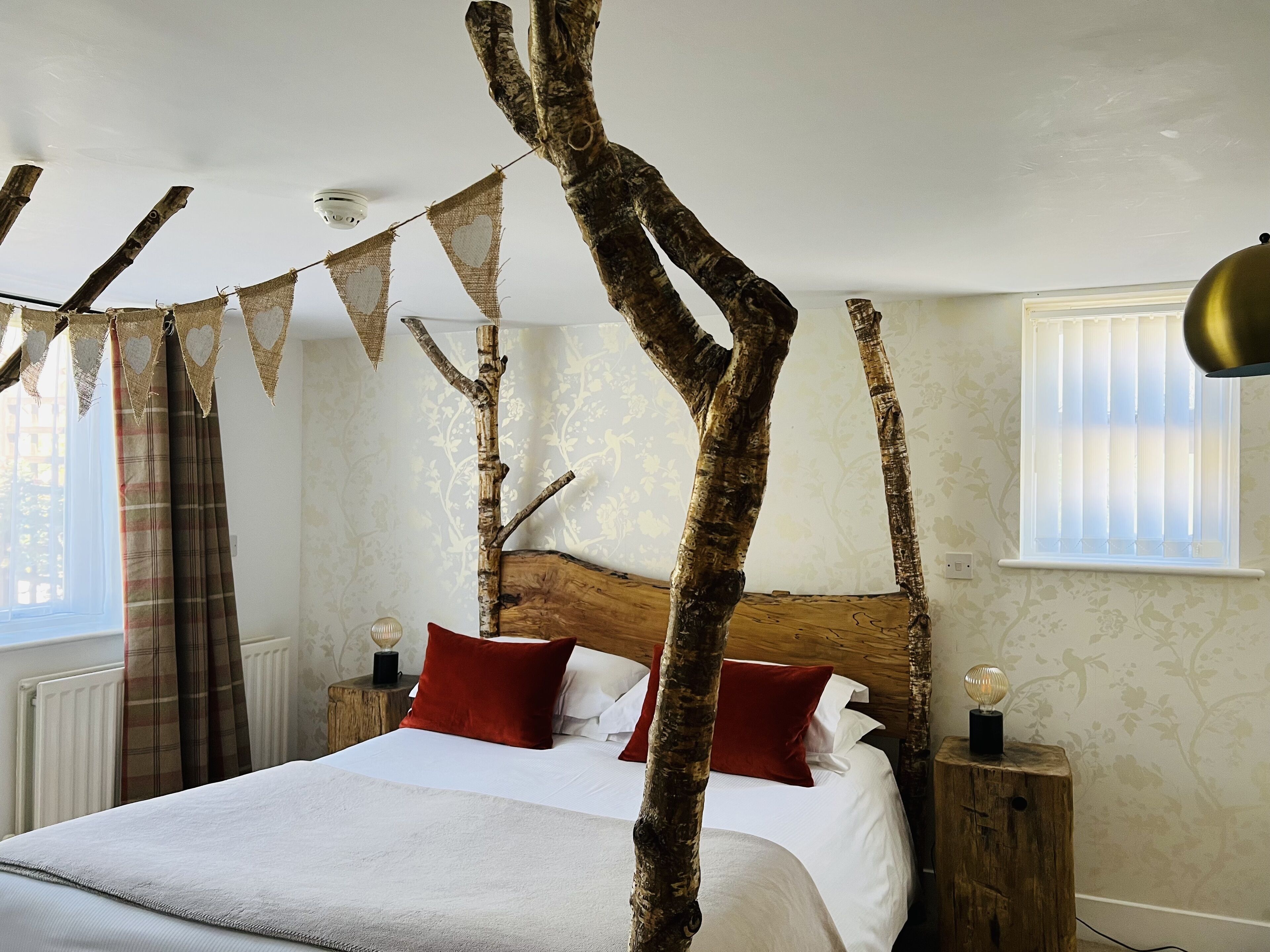 A bedroom with a bed, a wooden headboard, a window with blinds, and a wall with floral wallpaper.