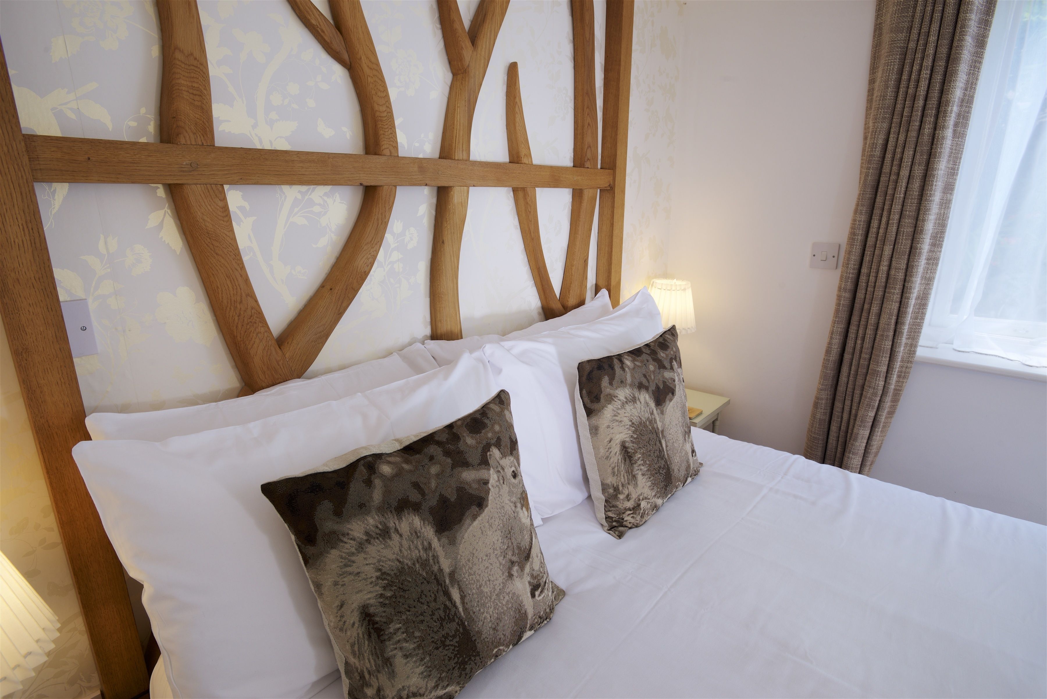 A neatly made bed with white bedding and two patterned pillows, a wooden headboard, a bedside lamp, and a window with curtains.