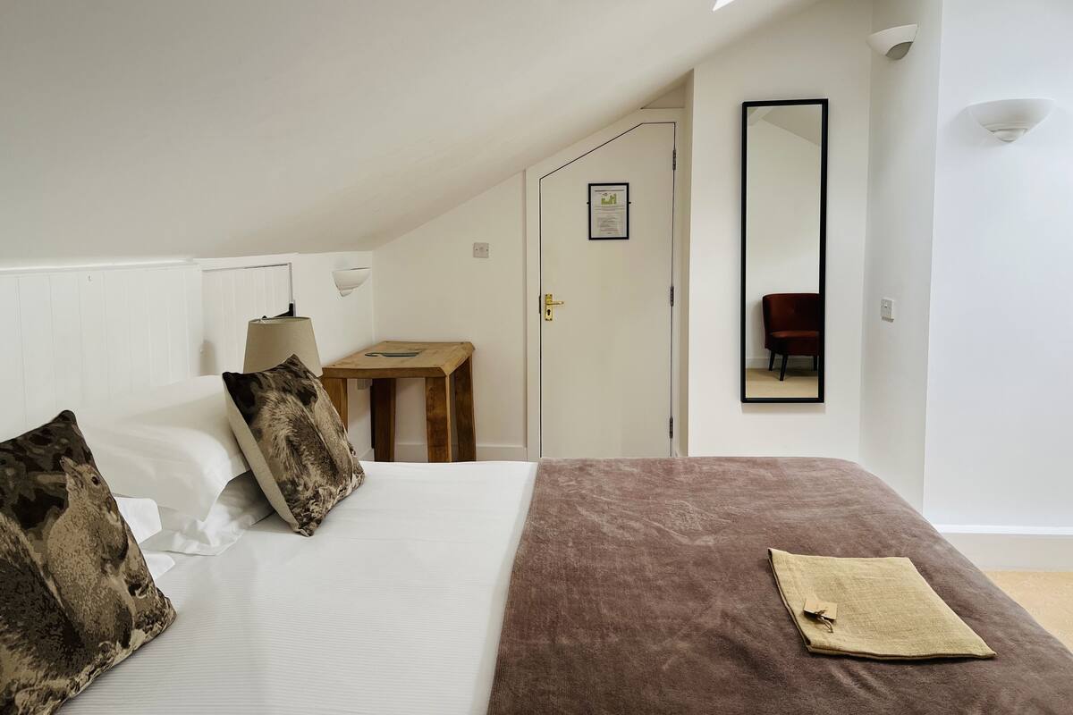 A bedroom with a sloped ceiling, a bed with a brown blanket, a small wooden table, a mirror, and a door leading to another room.