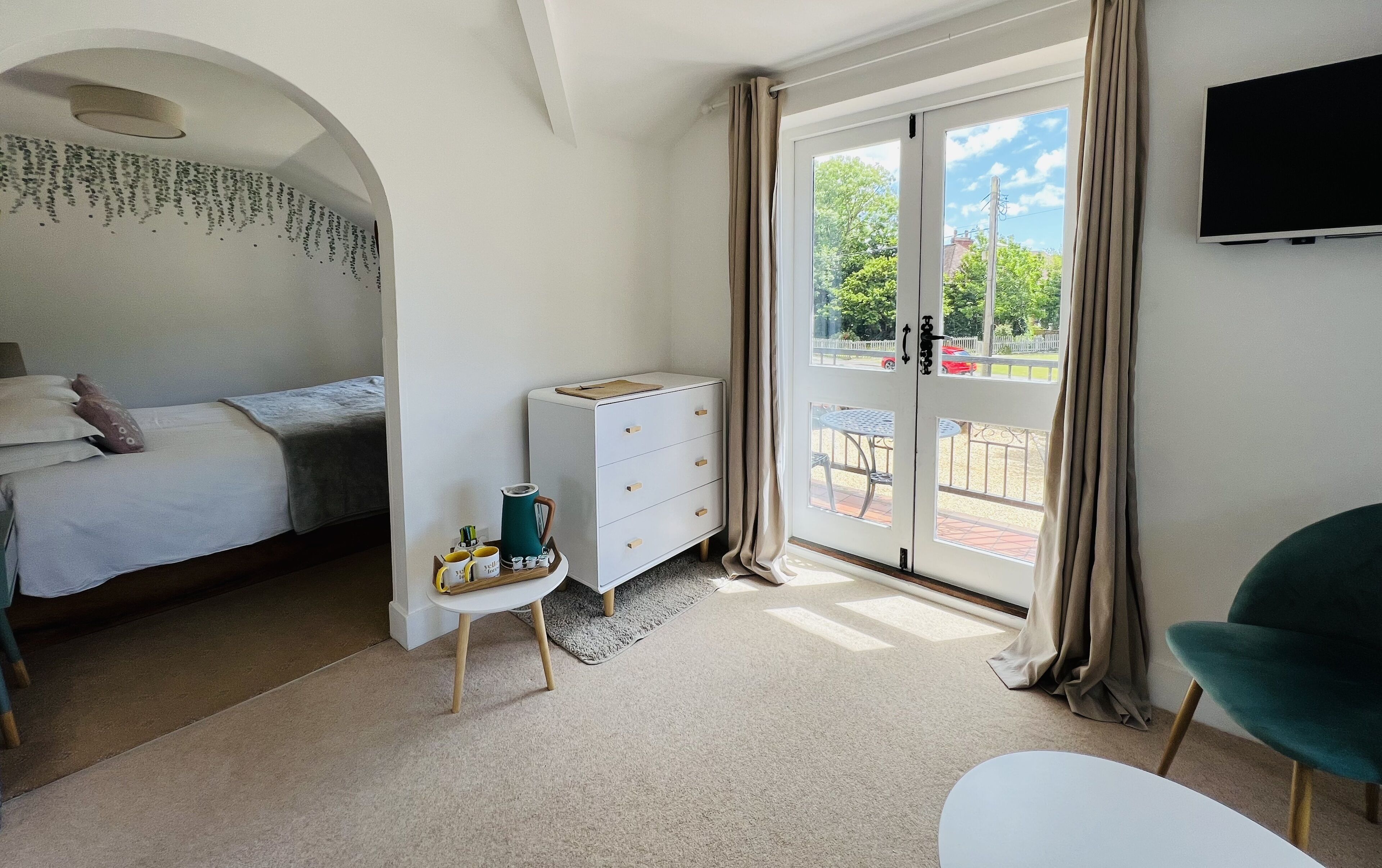 A bedroom with a bed, a dresser, a small table with items, and a view through a glass door to an outdoor area.