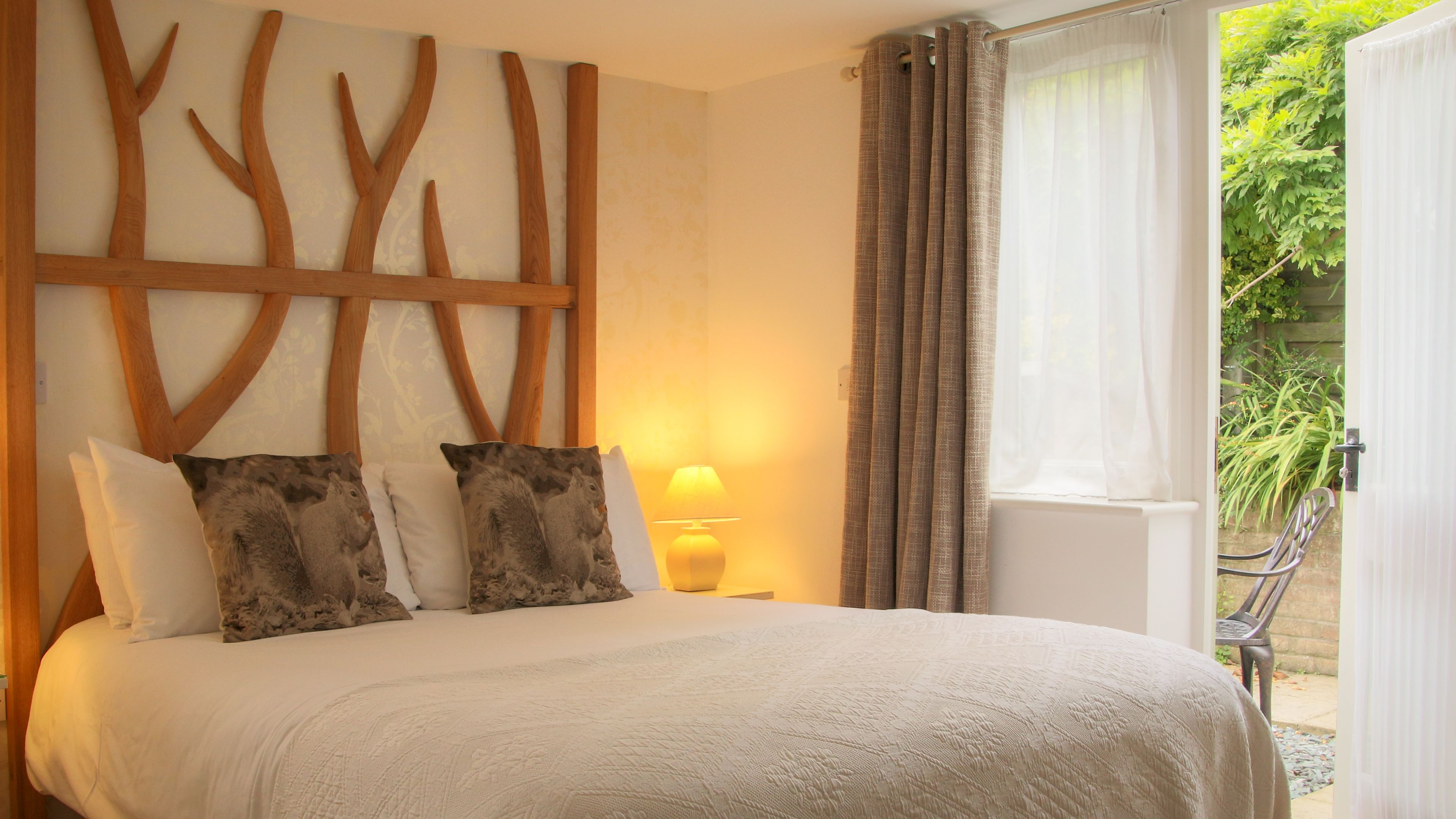 A bedroom with a bed, a wooden wall decoration, a lamp, and a view of greenery outside.